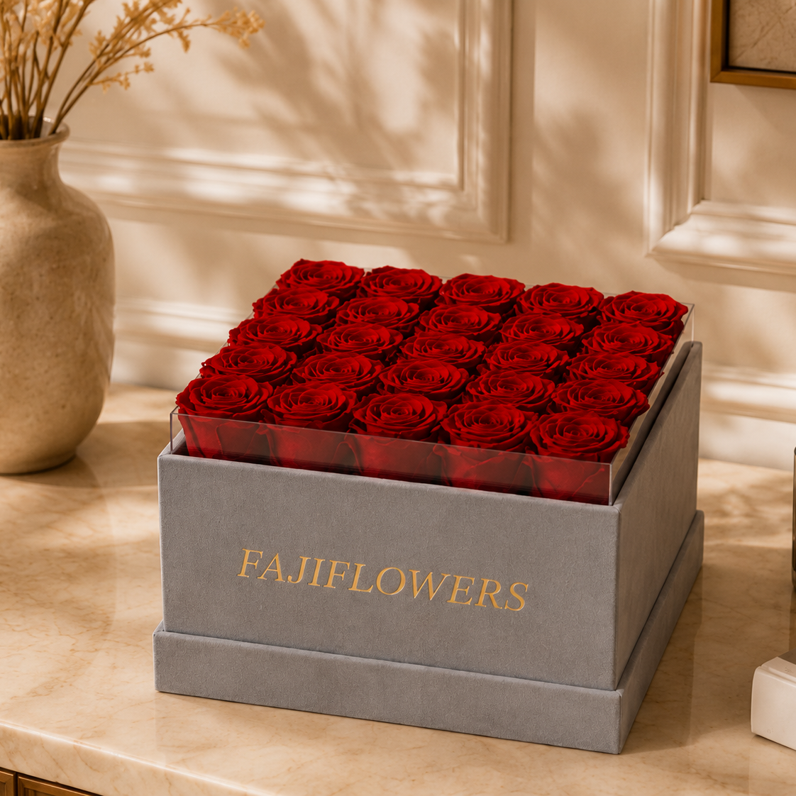 A square gray box of real preserved red roses with the brand name 'FAJIFLOWERS' in gold lettering on the front, displayed on a beige marble countertop in a warm, elegant interior setting.