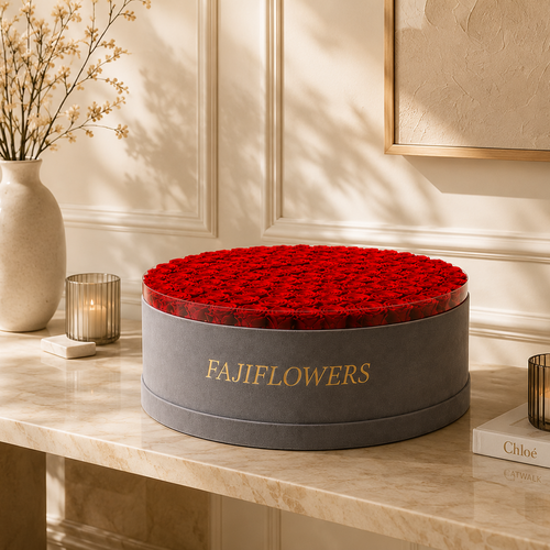 A round gray gift box filled with red roses sits on a marble console table in a warm, elegant room. The box is branded with gold lettering reading 'FAJIFLOWERS'.