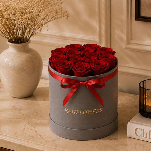 A round gray box filled with red roses, tied with a red ribbon and printed with the brand name 'FAJIFLOWERS', displayed on a marble surface next to a lit candle and a vase of dried flowers.