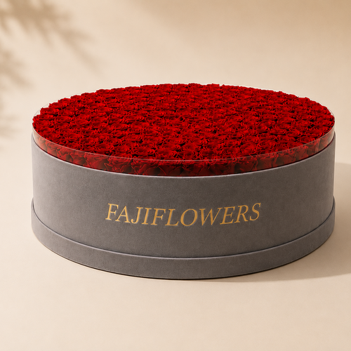 A round gray floral gift box with the brand name 'FAJIFLOWERS' in gold lettering, filled with a dense bed of deep red roses, set against a warm beige background with soft leaf shadows.