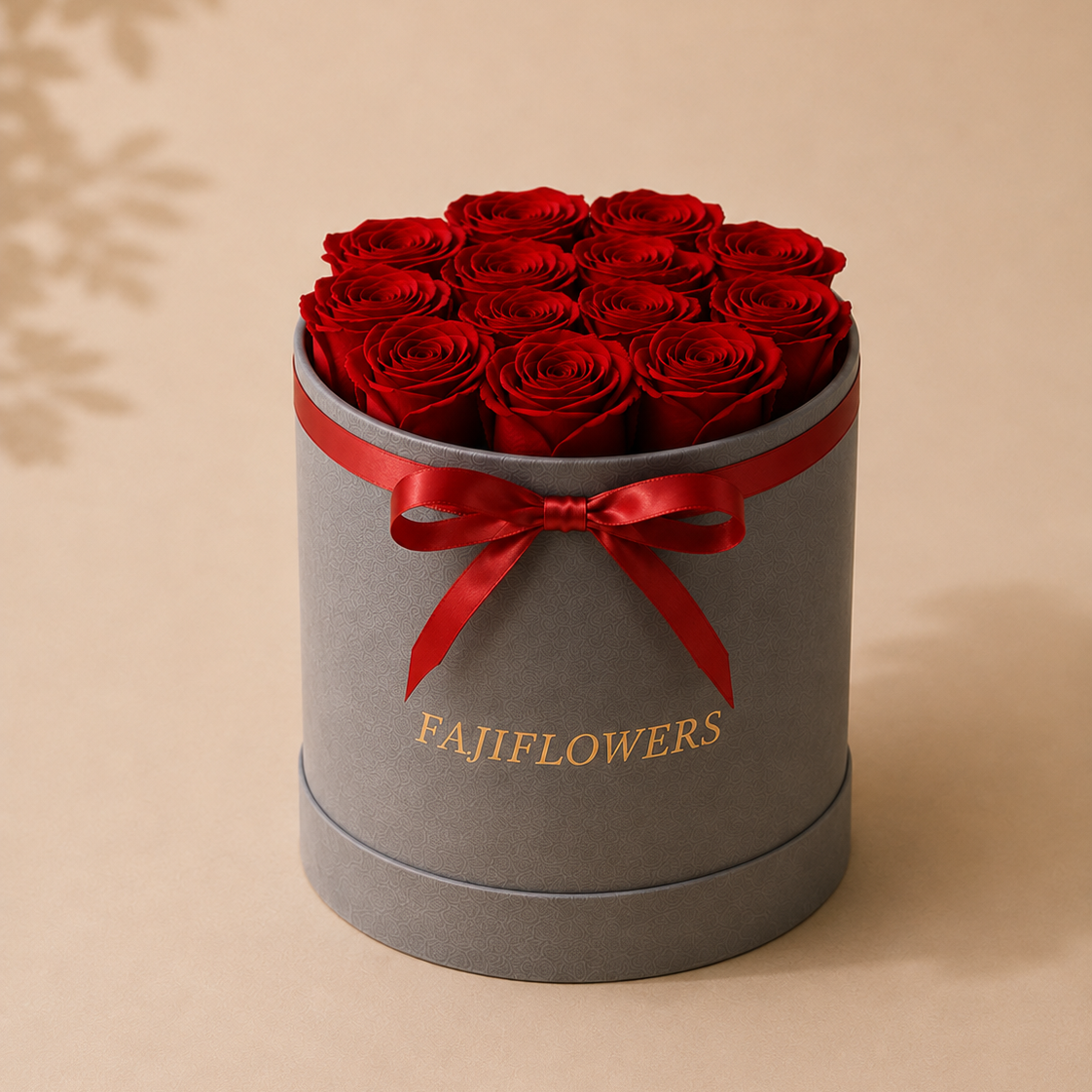 A round gray gift box filled with red roses, tied with a red ribbon and bow, with the brand name 'FAJIFLOWERS' printed in gold on the front, set against a warm beige background.