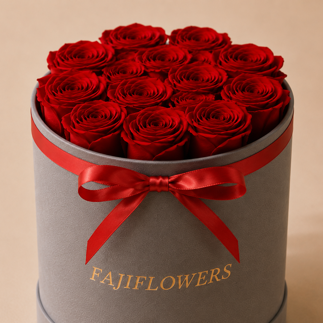 A round gray gift box filled with red roses, tied with a red satin ribbon and bow, with the brand name 'FAJIFLOWERS' printed in gold on the front, set against a solid beige background.