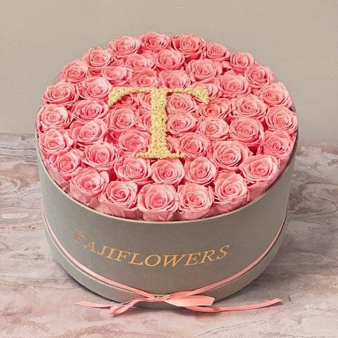 A decorative arrangement of real preserved red roses in a plush box with a custom initial on top, adorned with a bow with the letter 'T' on it.