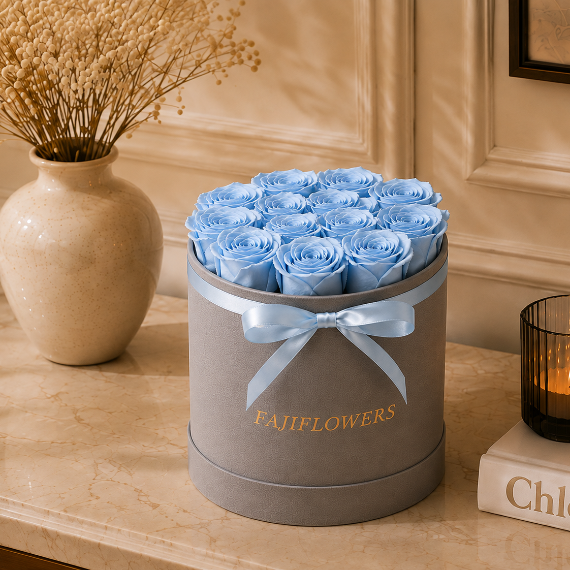 A round gray gift box filled with light blue roses, tied with a pale blue ribbon and printed with the word 'CAJIFLOWERS', displayed on a beige marble surface beside a vase of dried flowers and a lit candle.