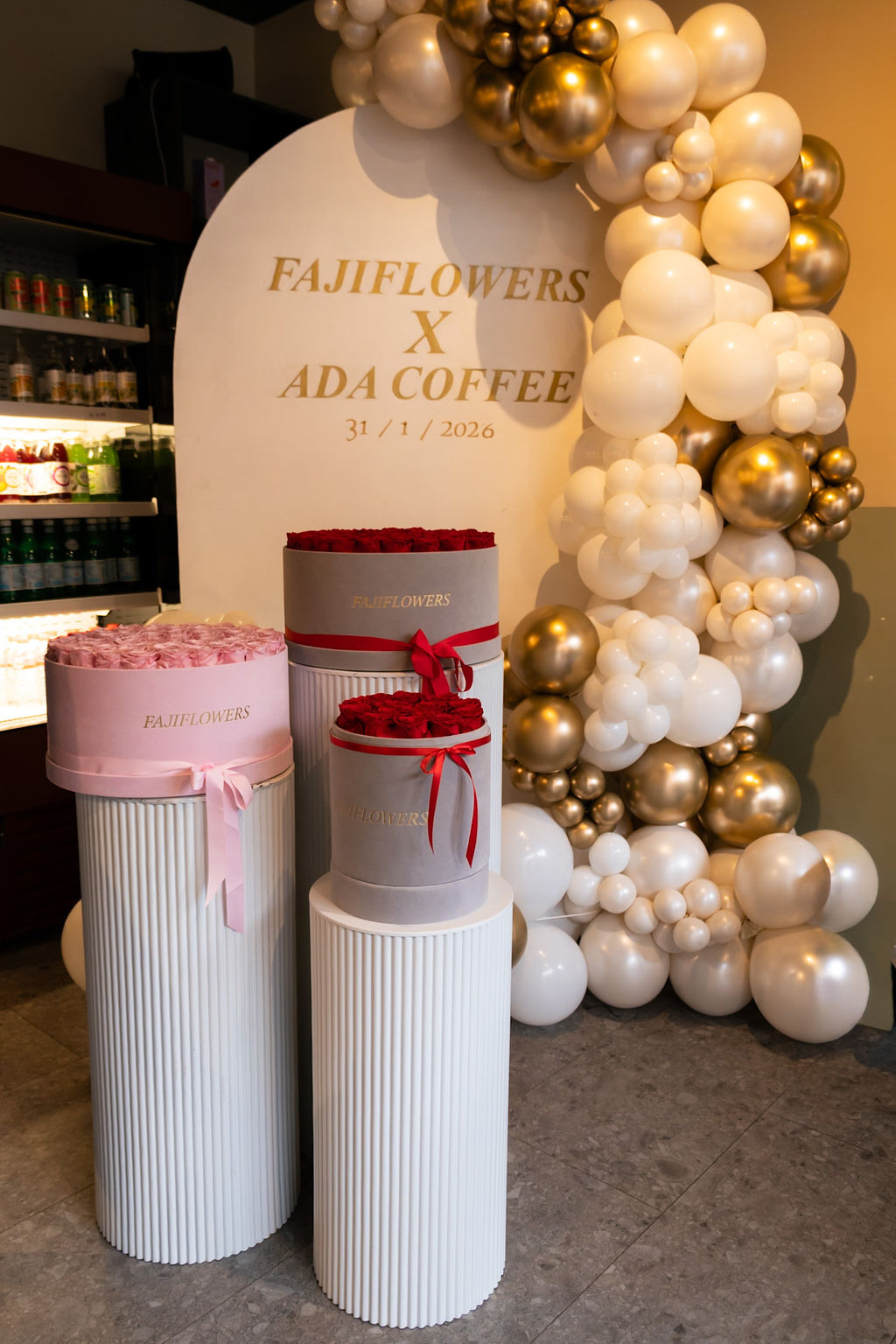 Pink and red FajiFlowers rose boxes displayed on white plinths beside balloons at Ada Coffee.