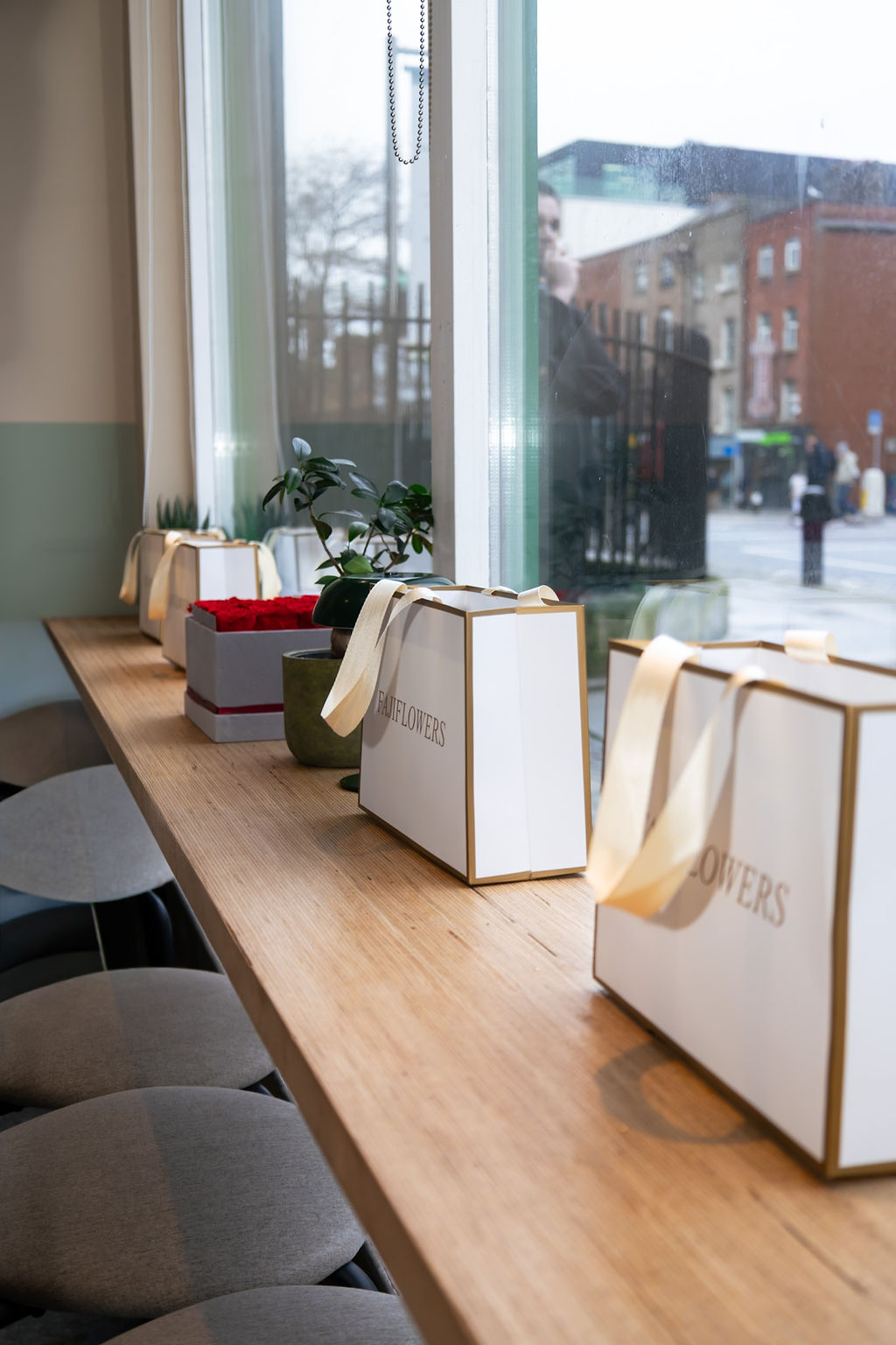 White FajiFlowers gift bags lined up on a café counter beside the window.